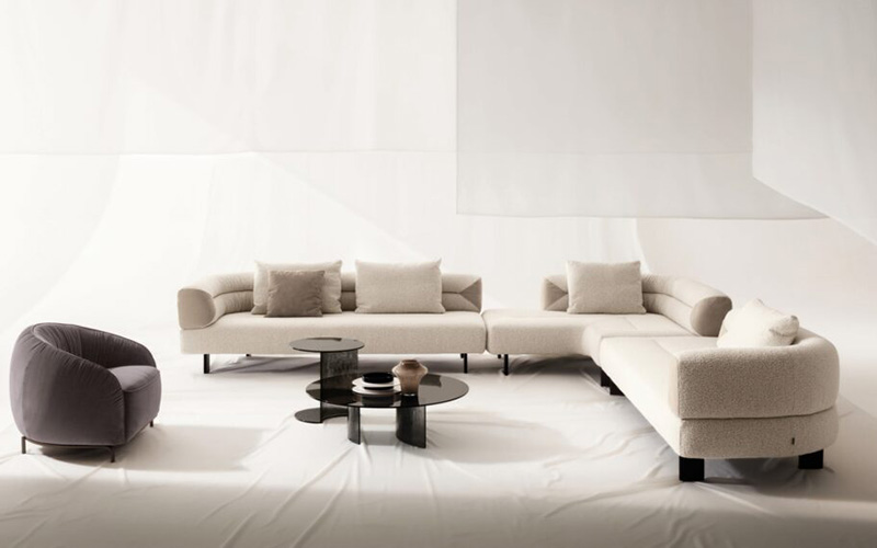 A modern living room featuring a large, curved cream sectional sofa, a dark gray armchair, and two tiered black coffee tables against a minimalist white backdrop.