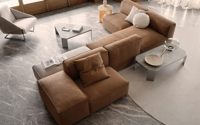 A modern living room with a modular brown sectional sofa arranged on a gray marble floor, paired with minimalist metal side tables, neutral cushions, and soft natural light from large windows.