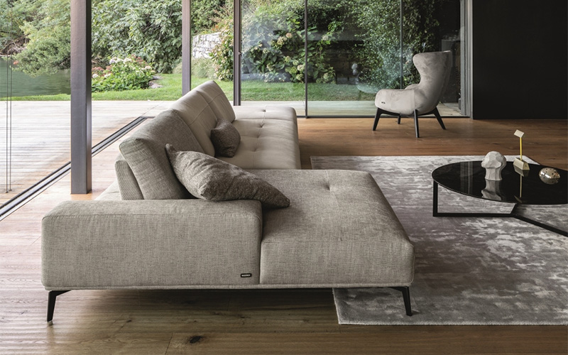 A wide, modern living room featuring a light grey modular sectional sofa, a black circular coffee table on a grey rug, and floor-to-ceiling glass doors opening onto an outdoor deck.