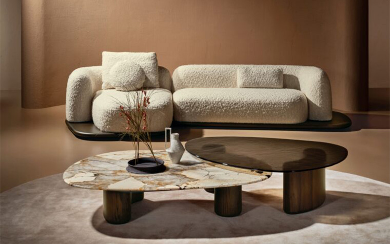 Contemporary living room with a curved cream sofa, textured upholstery, and sculptural marble and wood coffee tables in warm neutral tones.